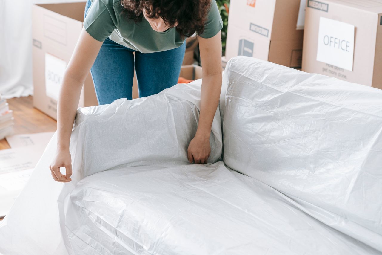 gallery-02 A woman is carefully covering a sofa with protective wrapping while packing boxes in a bright room.